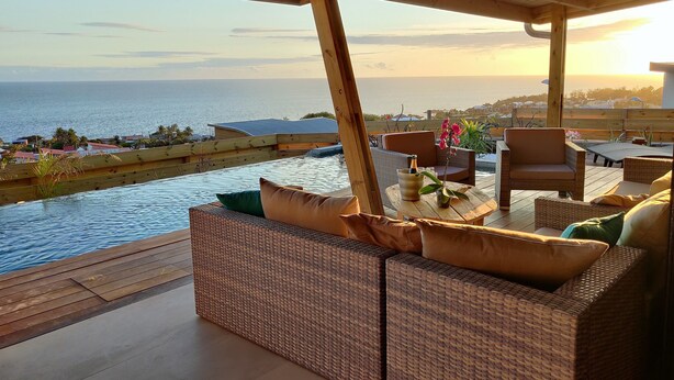 Une terrasse en bois avec des meubles en osier, une table ornée d’une bouteille et de fleurs, et une vue sur la mer et les bâtiments en arrière-plan.