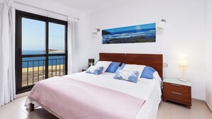 Un dormitorio con una cama grande, una mesita de noche, una lámpara de pared y un cuadro de una playa.