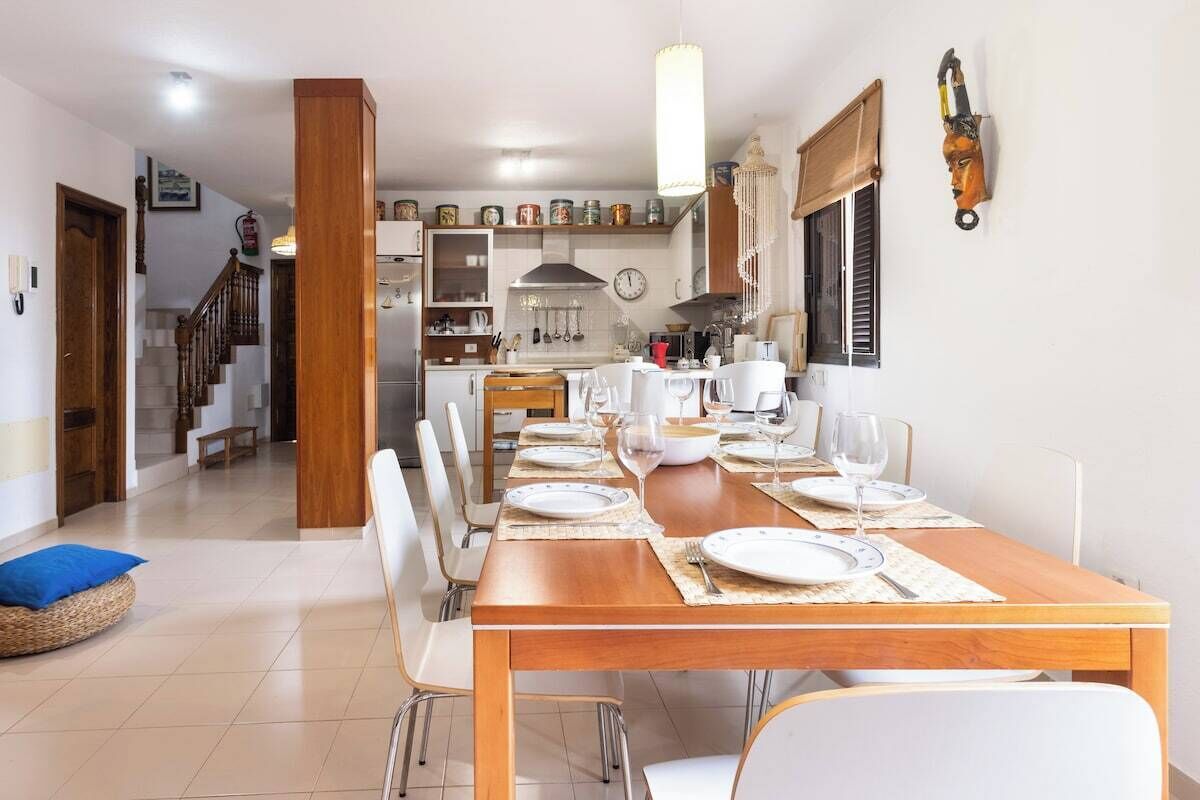 Un comedor con una mesa de madera dispuesta para cuatro personas, sillas blancas y una cocina al fondo.