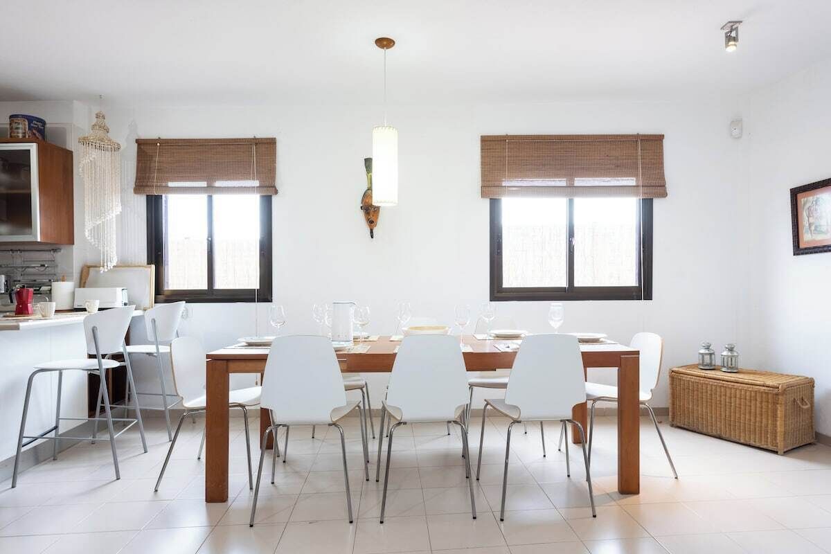 Une cuisine moderne avec des meubles blancs, une table à manger avec des chaises et deux fenêtres équipées de stores bruns.
