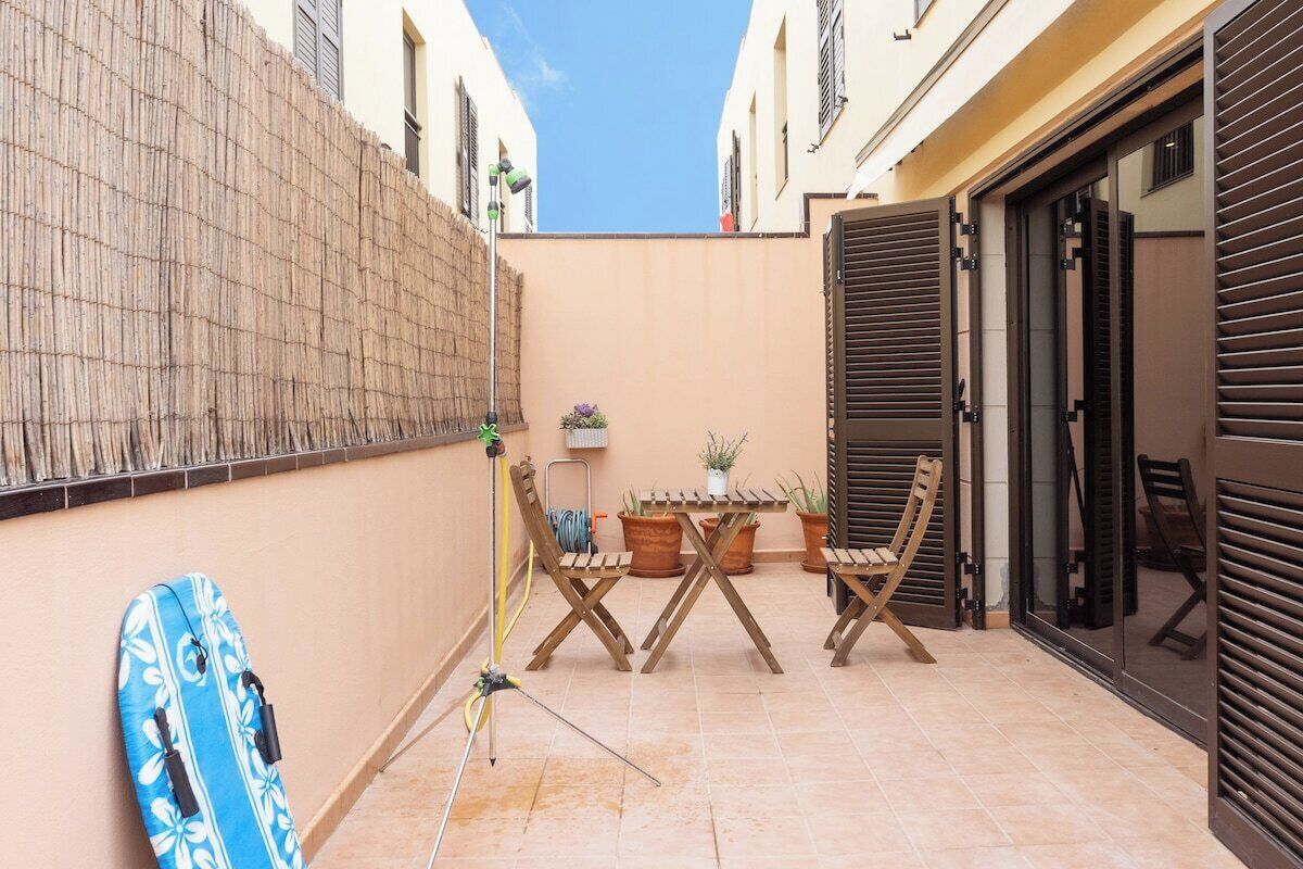 A narrow courtyard with a bamboo wall, a folding table, two chairs, a surfboard, and a potted plant.