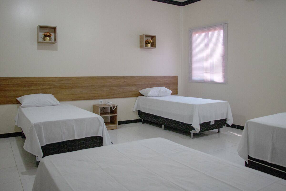 A hotel room with three single beds, a wooden headboard, a bedside table with a lamp, and a window with blinds.
