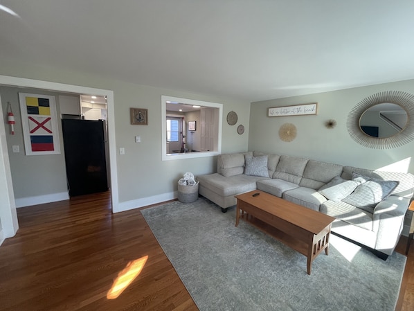A living room with a grey sectional sofa, a wooden coffee table, and a grey rug. There is a window providing natural light and a door leading to another room.