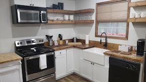 Eine moderne Küche mit weißen Schränken, einem Edelstahlherd, einer Mikrowelle und einem Geschirrspüler. Darüber befinden sich Holzregale und ein Fenster mit Jalousien.