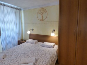 A hotel room with a bed, two bedside lamps, a nightstand, a wooden wardrobe, and a wall with a tree design. - TIERRA - Apartments Bevk in charming Piran (4 Piran)