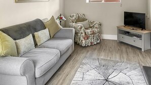 Un salon avec un canapé gris, un fauteuil à motifs floraux, un meuble pour la télévision et une grande fenêtre donnant sur un beau paysage.