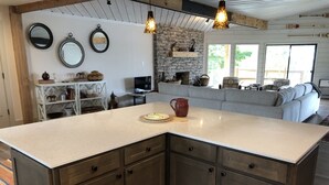 Een keuken met een groot eiland, houten kasten en een stenen muur. Er zijn drie hangende lampen, een open haard en een zithoek met een bank.