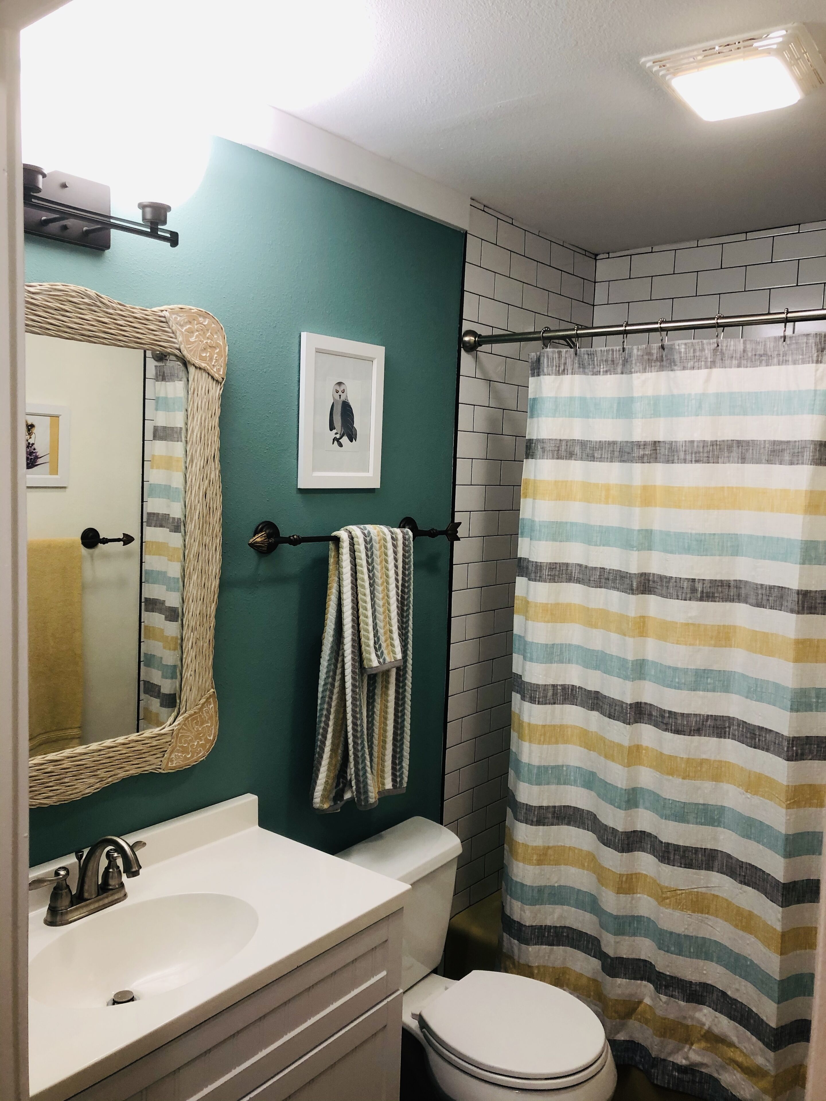 A bathroom with a white sink, a toilet, a striped shower curtain, and a mirror with a woven frame.