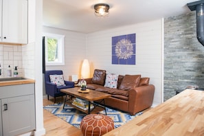 Ein modernes Wohnzimmer mit einer braunen Ledersofa, einem Holztisch und einem blau gemusterten Teppich. Ein Kamin aus Stein, ein Fenster mit Blick auf Grünflächen und eine Wandleuchte sind ebenfalls vorhanden.
