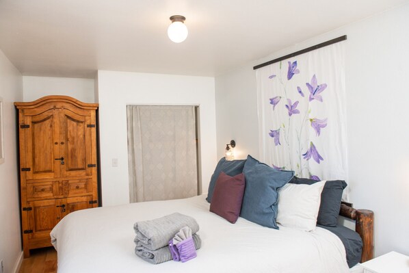 Un dormitorio con cama, almohadas, un armario de madera, una ventana con cortinas y una lámpara fijada a la pared.