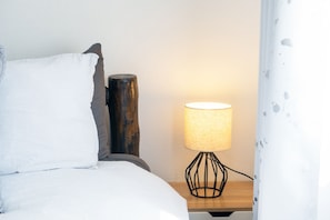 Ein Bett mit weißer Bettwäsche, ein Nachttisch aus Holz mit einer Lampe und ein teilweise sichtbarer weißer Vorhang.