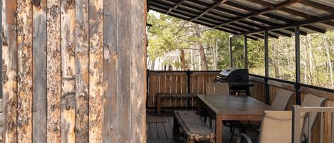 Eine Terrasse im Freien mit Holztisch und -stühlen, einem Grill und einer Metall-Dachkonstruktion.