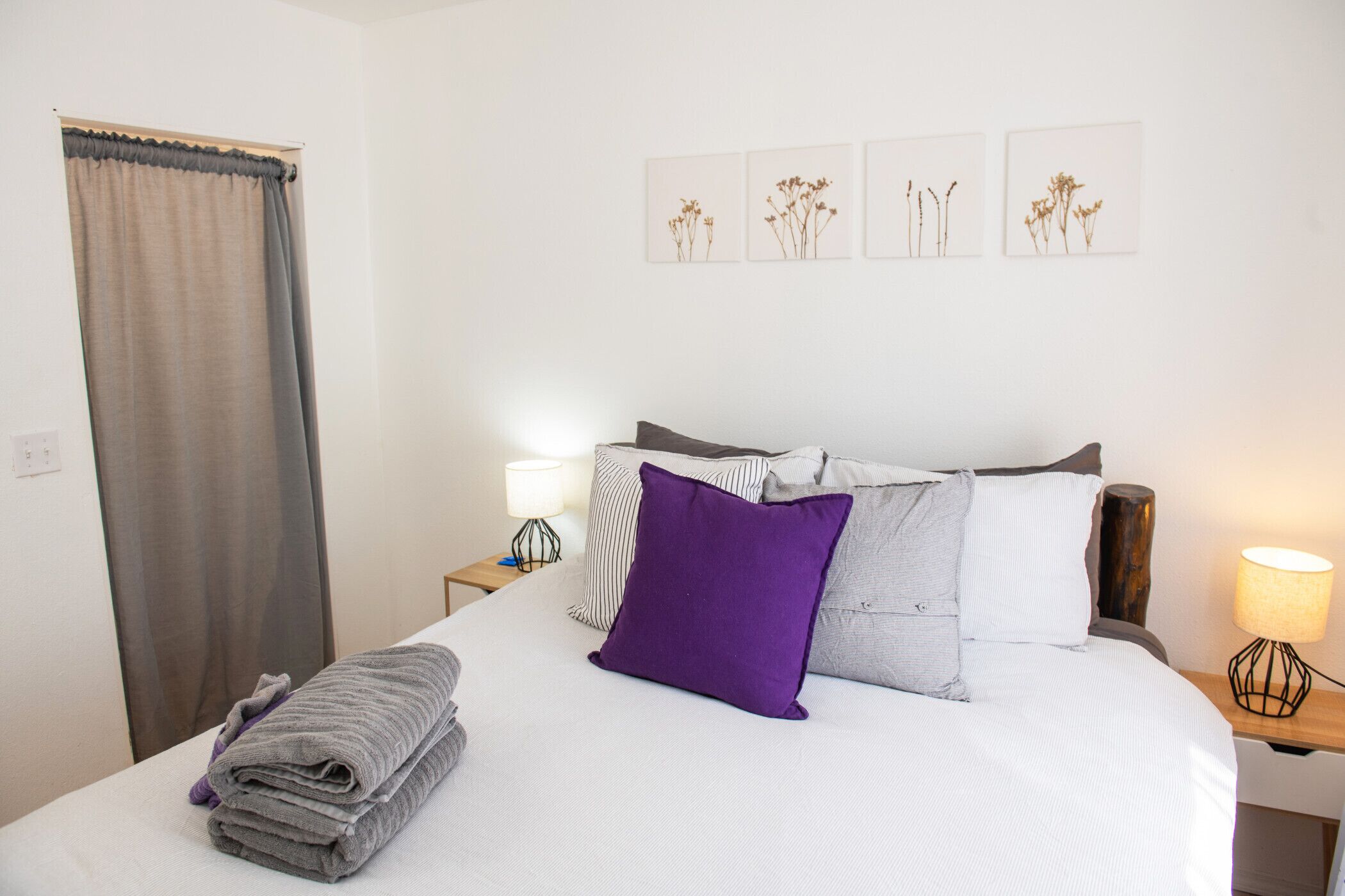 A neatly made bed with a white sheet, a purple pillow, and a folded gray towel. There are two bedside tables with lamps, a curtain, and wall art depicting plants.