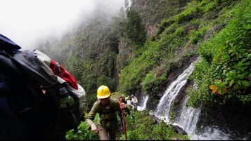 Une personne portant un casque jaune et des vêtements de camouflage, avec un sac à dos, marche près d’une cascade dans une zone forestière verdoyante et luxuriante.