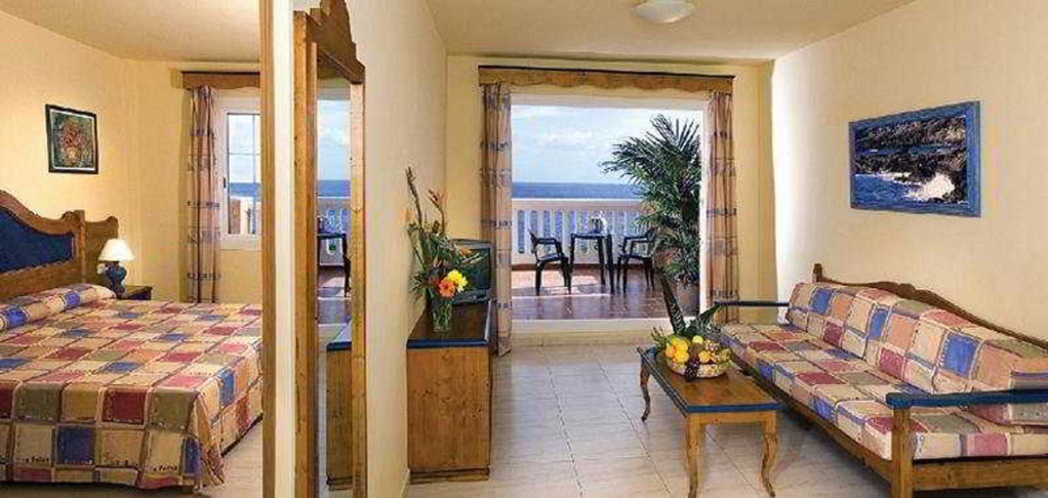 Ein Hotelzimmer mit Bett, einer Couch, einem kleinen Tisch mit einer Vase und einem Balkon mit Meerblick.