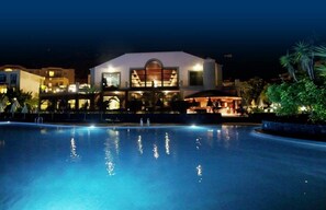 Vista nocturna de un hotel con piscina, zona de bar y un edificio grande con un ventanal arqueado destacado.
