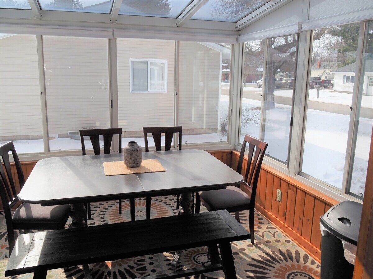 Een glazen orangerie met een tafel, stoelen, grote ramen en uitzicht op een woning.