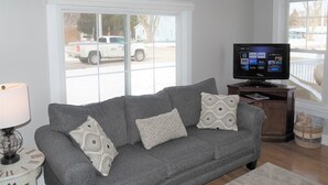 Una sala de estar con un sofá gris, cojines estampados, un televisor de pantalla plana y una ventana que ofrece vistas a un coche aparcado y una casa.