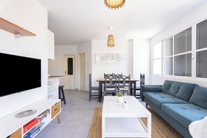 A modern living room with a flat-screen TV, a white coffee table, a blue sofa, and a dining area with a table and chairs.