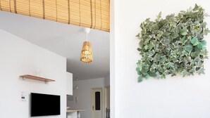 Una sala de estar moderna con televisor, un sillón de mimbre con cojín de lunares, una mesita con planta y un adorno de follaje montado en la pared.