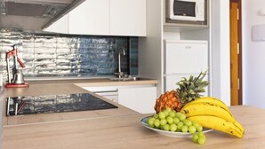 Una cocina moderna con armarios blancos, un lavamanos de acero inoxidable y un microondas. En primer plano, una mesa de madera con un plato de fruta.