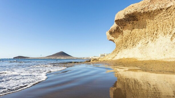 Una playa con una gran formación rocosa y una montaña al fondo.