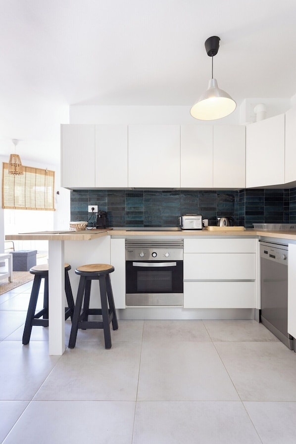 Una cocina moderna con armarios blancos, horno empotrado, lavavajillas y encimera de madera. Hay dos taburetes en la isla de la cocina.