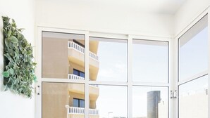 Habitación de esquina con una silla de mimbre, una mesita redonda con una planta en maceta, un ventanal con vista a un edificio y un tapiz decorativo.