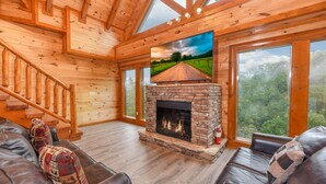 L’intérieur chaleureux d’un chalet avec une cheminée en pierre, des canapés en cuir, un escalier en bois et de grandes fenêtres offrant une vue sur l’extérieur.