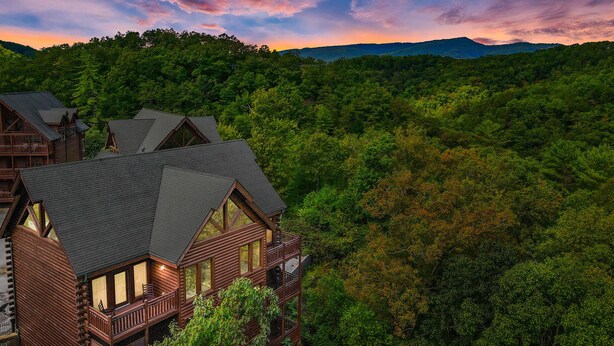 Une cabane en rondins à plusieurs niveaux, dotée de grandes fenêtres, entourée d’une forêt dense et avec des montagnes en arrière-plan.