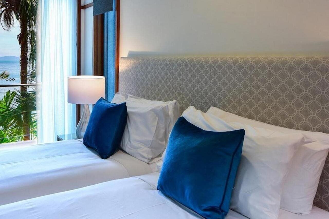 Ein Hotelzimmer mit Bett, Nachttisch, Lampe und Blick auf das Meer durch große Fenster.