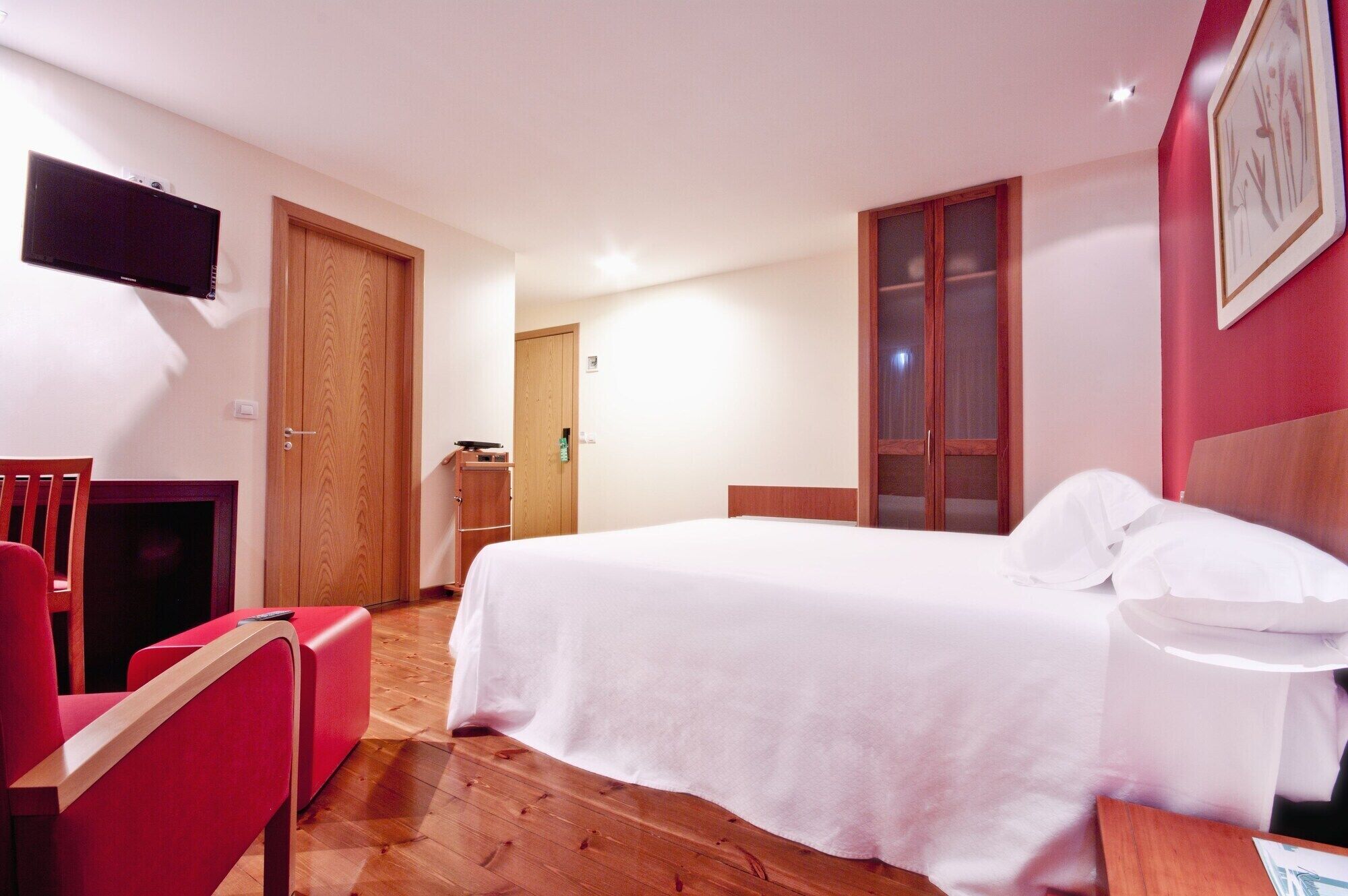 Habitación de hotel con cama, televisión montada en la pared, una silla roja y un armario de madera.