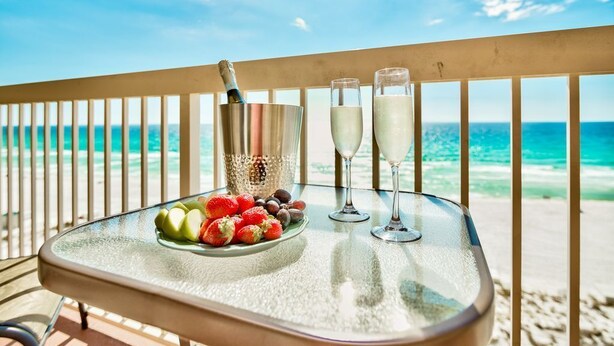 A glass table with a bottle of champagne, two glasses of champagne, and a plate of fruit on a balcony overlooking the beach. - Pelican Beach Resort, Beachfront 7th floor , White Beach, Pool, WiFi, Cable TV (Destin)