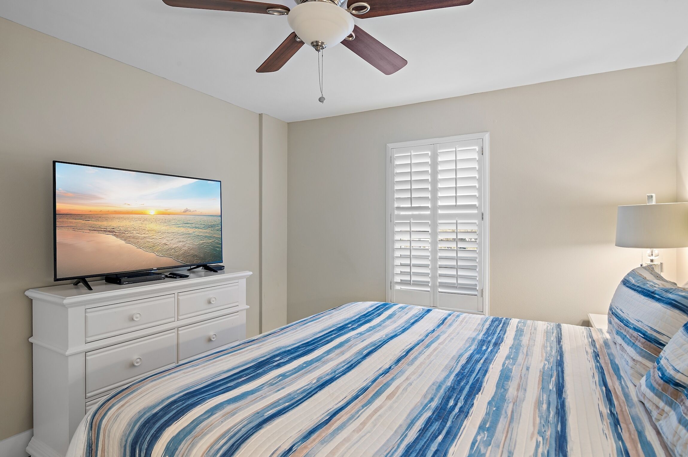 A bedroom with a bed, a TV on a dresser, a ceiling fan, and a window with blinds.