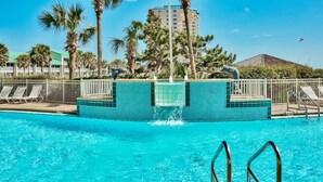 A swimming pool with a water fountain, surrounded by palm trees and a tall building in the background. - Pelican Beach Resort, Beachfront 7th floor , White Beach, Pool, WiFi, Cable TV (Destin)