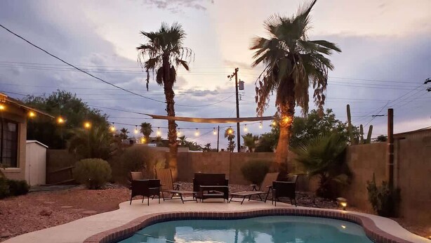 A swimming pool with a curved edge, surrounded by palm trees and outdoor furniture. - Phoenix Home | Pool & String Lights (Phoenix)