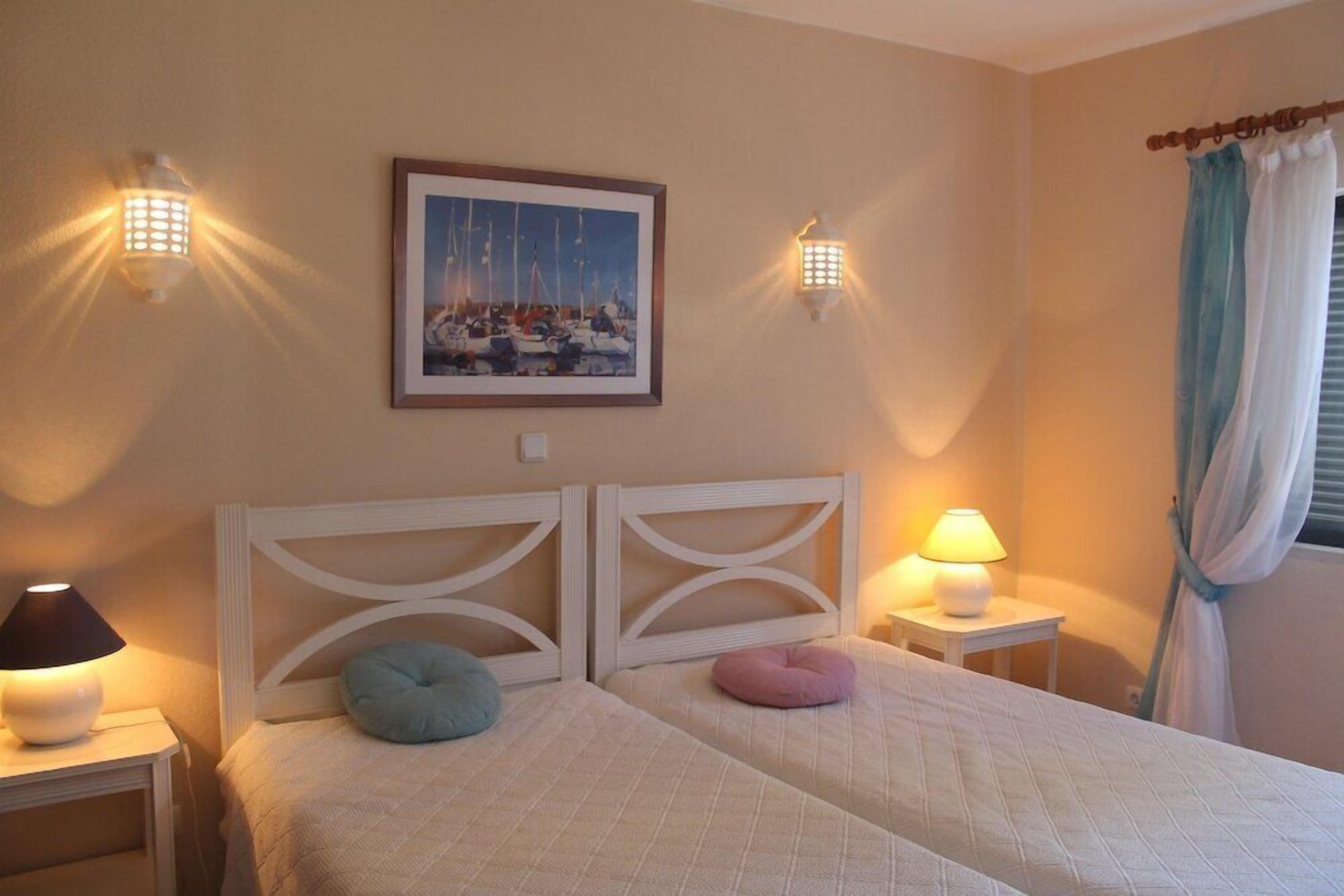 A hotel room with two beds, a framed picture on the wall, and two wall-mounted lamps.