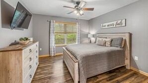 Een slaapkamer met een bed, een dressoir, een plafondventilator, een aan de muur bevestigde televisie en een raam met gordijnen.