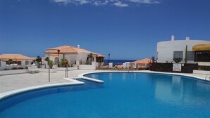 A swimming pool with a curved edge, surrounded by buildings with terracotta roofs, and a clear blue sky.
