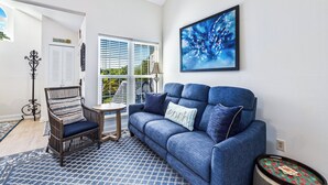 Uma sala de estar com sofá azul, tapete estampado, cadeira de madeira e janela com persianas.