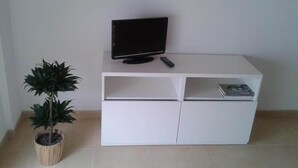 Un meuble blanc pour téléviseur, sur lequel est posé un téléviseur à écran plat et une télécommande, une petite plante en pot et une étagère avec un livre et un lecteur DVD.