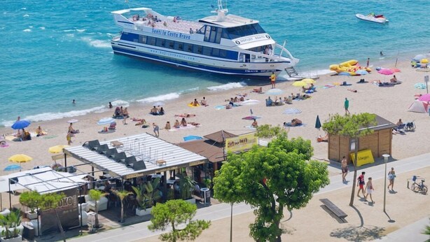 A beachside view with a large boat named 'Senti la Costa Bravo' and a beach area with umbrellas, sun loungers, and a small wooden structure.