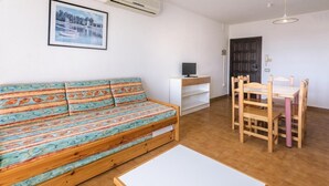 A furnished room with a sofa, a small table, a TV, and a dining area with chairs.