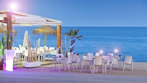 An outdoor seating area with white chairs and tables, a thatched umbrella, and a view of the sea.