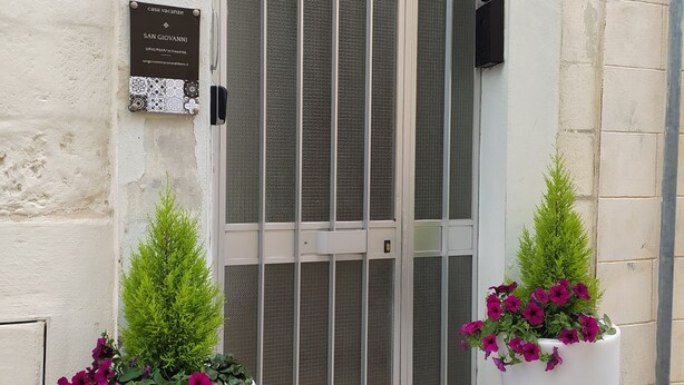 A building entrance with a metal gate, a plaque, and two large flower pots. - Vacation home San Giovanni nei Sassi (Matera)