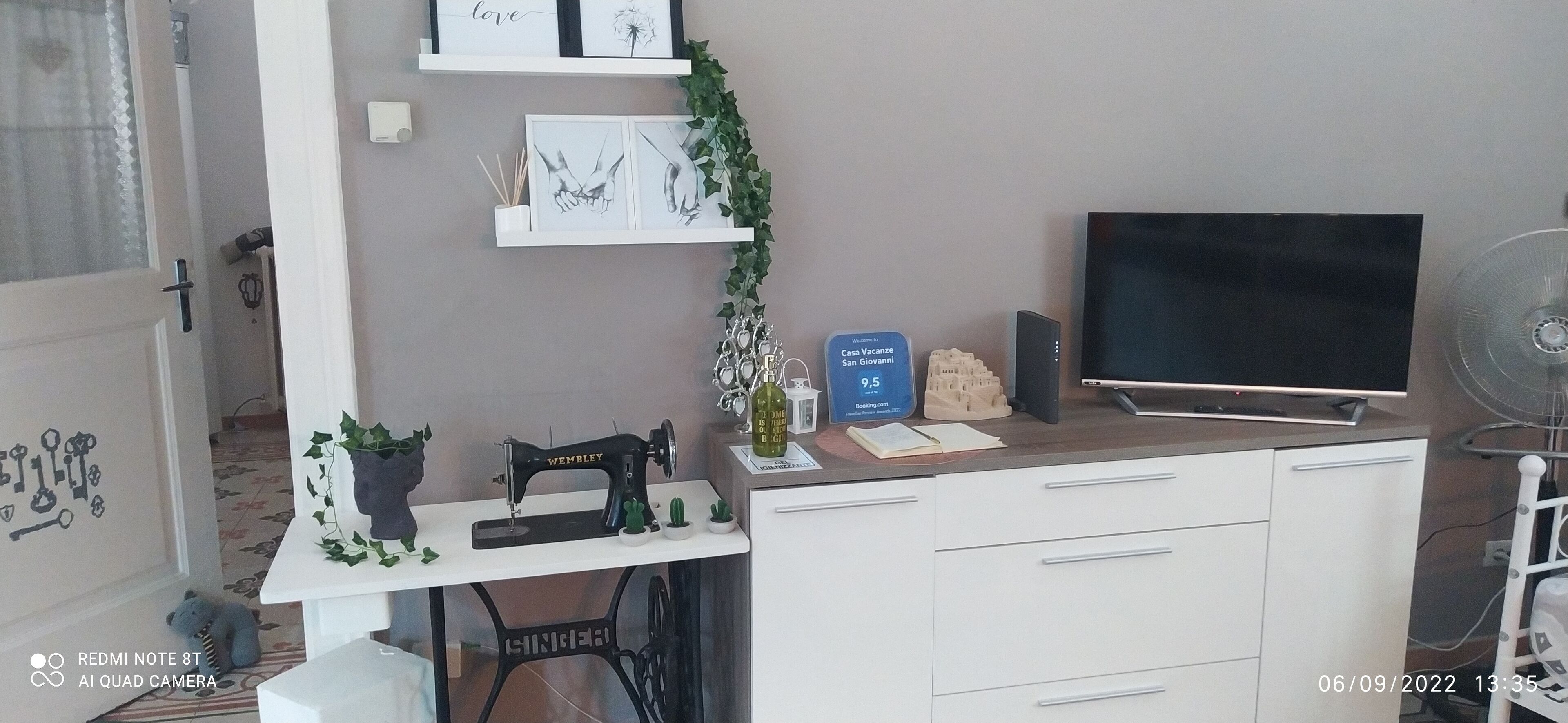 A room with a sewing machine, a flat-screen TV, a small table with a plant, and a sign indicating a room number.