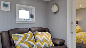 Une pièce avec un canapé en cuir marron, deux coussins décoratifs à motifs, une horloge murale, une fenêtre avec une plante et un lit recouvert d’une couvre-lit à fleurs.