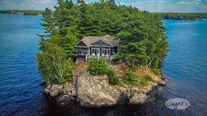 A house on a small island surrounded by water, with a dock and a large rock in the foreground.