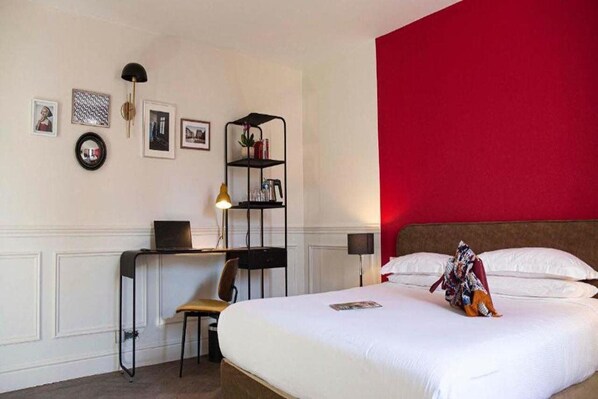 Una habitación de hotel con una cama, un escritorio con silla, una estantería y una pared decorada en rojo.