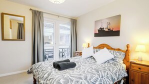 A bedroom with a bed, a nightstand, a lamp, a mirror, a window with curtains, and a painting on the wall. - 3 bedroom accommodation in Brancaster Staithe, near Wells-next-the-Sea (Brancaster Staithe, near Wells-next-the-Sea)